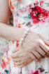 A close-up of a woman wearing a white sleeveless dress with a vibrant red, pink, and orange floral print. She is wearing a pearl bracelet on her wrist, with her hands crossed over her waist, set against a blurred brick wall background.