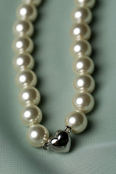 A close-up of a pearl necklace with a polished silver heart-shaped charm, resting on a textured light green fabric background.