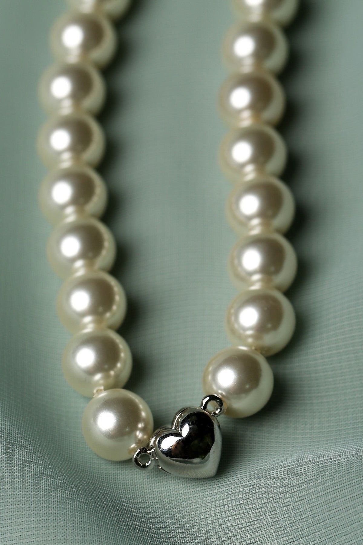 A close-up of a pearl necklace with a polished silver heart-shaped charm, resting on a textured light green fabric background.