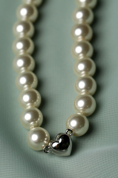 A close-up of a pearl necklace with a polished silver heart-shaped charm, resting on a textured light green fabric background.