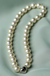 A strand of white freshwater pearls arranged in a loop on a light sage green satin fabric background, featuring a small silver heart-shaped charm at the end.