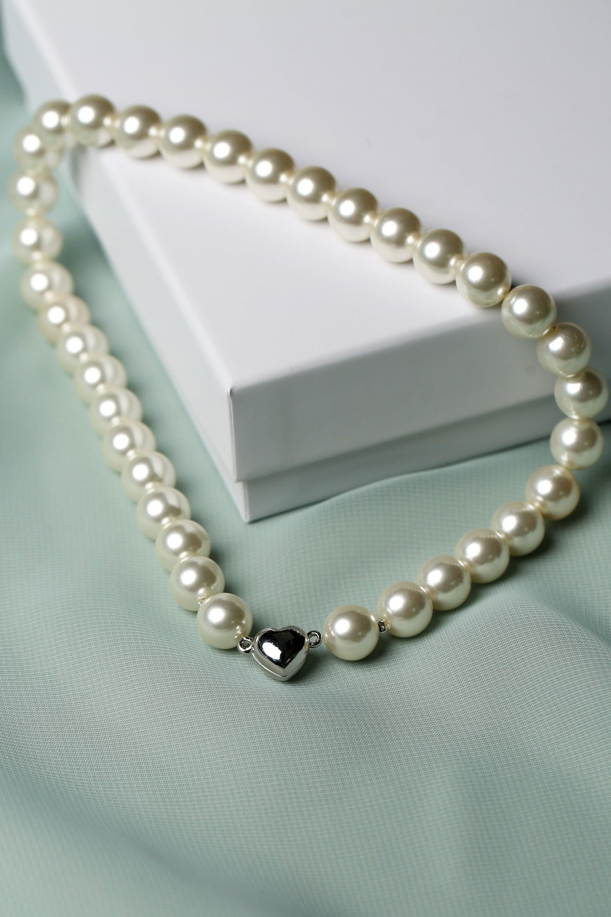 A strand of cream-colored freshwater pearls with a polished silver heart-shaped charm, draped elegantly over a white gift box on a soft mint green fabric background.