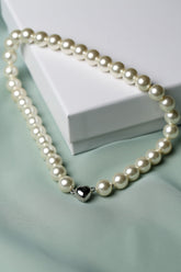 A strand of cream-colored freshwater pearls with a polished silver heart-shaped charm, draped elegantly over a white gift box on a soft mint green fabric background.