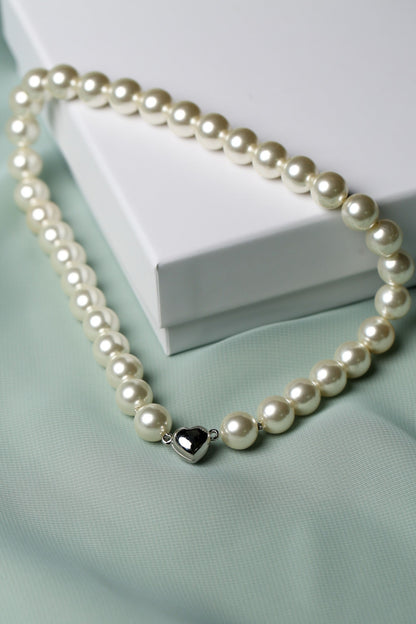 A strand of cream-colored freshwater pearls with a polished silver heart-shaped charm, draped elegantly over a white gift box on a soft mint green fabric background.