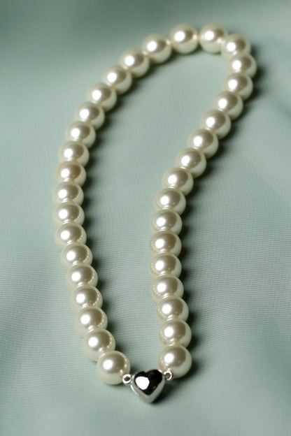 A strand of white pearl-like beads arranged in a loop on a soft mint green fabric background, with a small silver heart charm at the center.