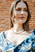 A woman wearing a blue floral dress with a pearl necklace, standing outdoors against a red brick wall.