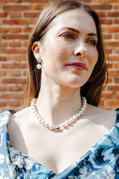 A woman wearing a blue floral off-the-shoulder dress and a pearl necklace, standing in front of a red brick wall.