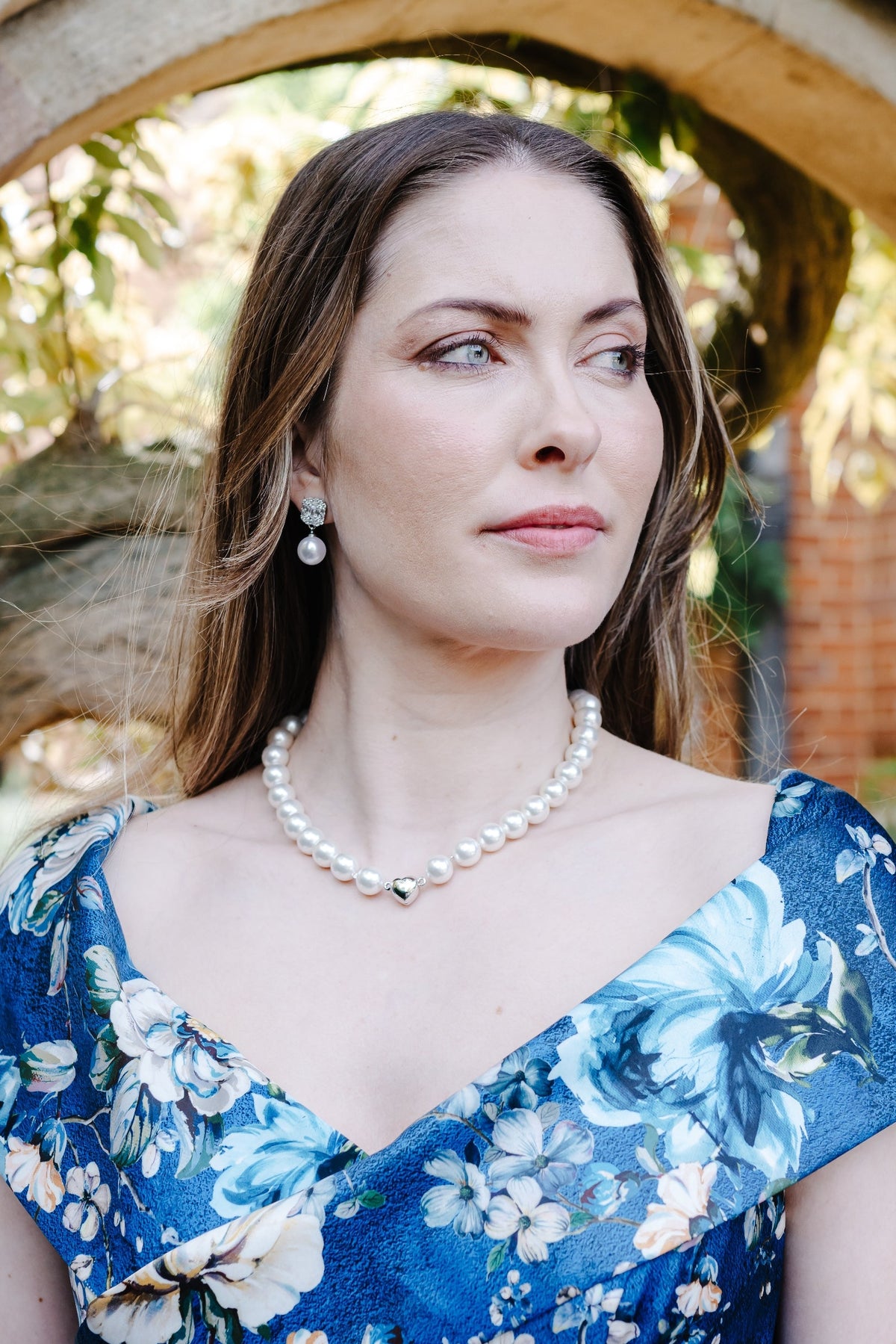 A woman wearing a blue floral off-the-shoulder dress and a pearl necklace stands framed by a stone archway in a garden setting.