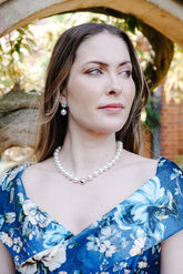 A woman wearing a blue floral off-the-shoulder dress and a pearl necklace stands framed by a stone archway in a garden setting.