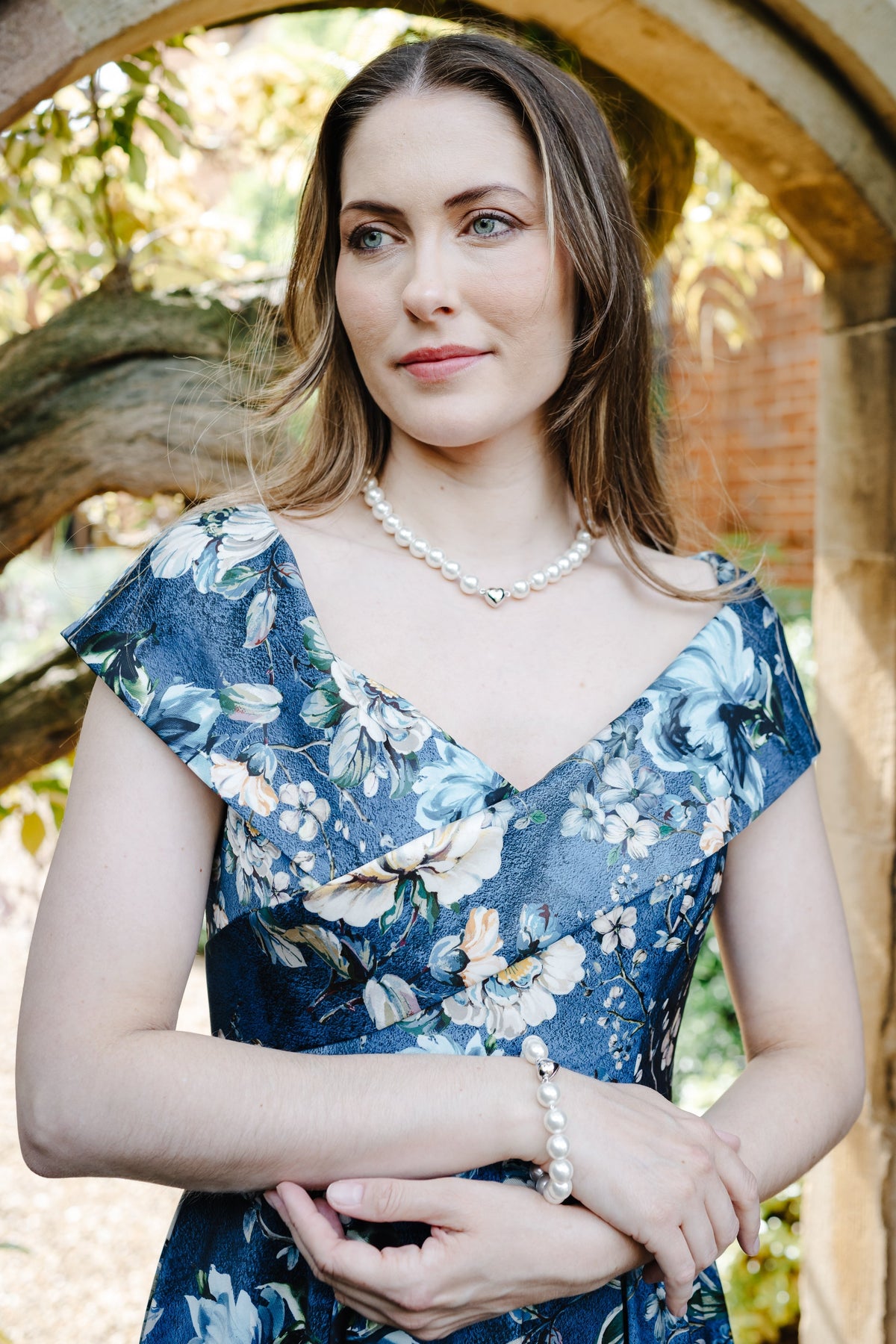 A woman wearing a blue floral dress and pearl jewelry stands framed by a stone archway in an outdoor garden setting.