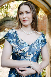 A woman wearing a blue floral dress and pearl jewelry stands framed by a stone archway in an outdoor garden setting.