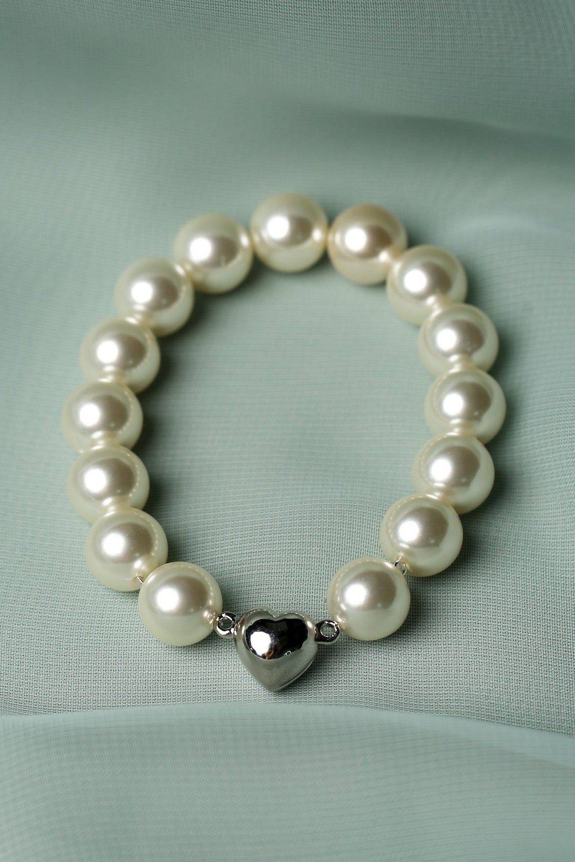 A pearl beaded bracelet with a polished silver heart charm, displayed on a draped mint green fabric background.