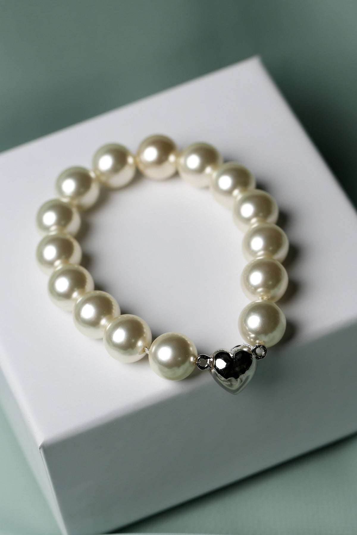 A pearl beaded bracelet with a polished silver heart charm, displayed on a white gift box against a soft sage green fabric background.