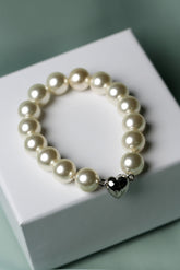A pearl beaded bracelet with a polished silver heart charm, displayed on a white gift box against a soft sage green fabric background.