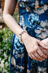 A close-up lifestyle shot of a person wearing a blue floral dress and a beaded pearl bracelet, standing outdoors with green foliage in the background.