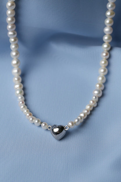 A pearl necklace with a polished silver heart pendant, displayed on a soft, draped light blue fabric background.
