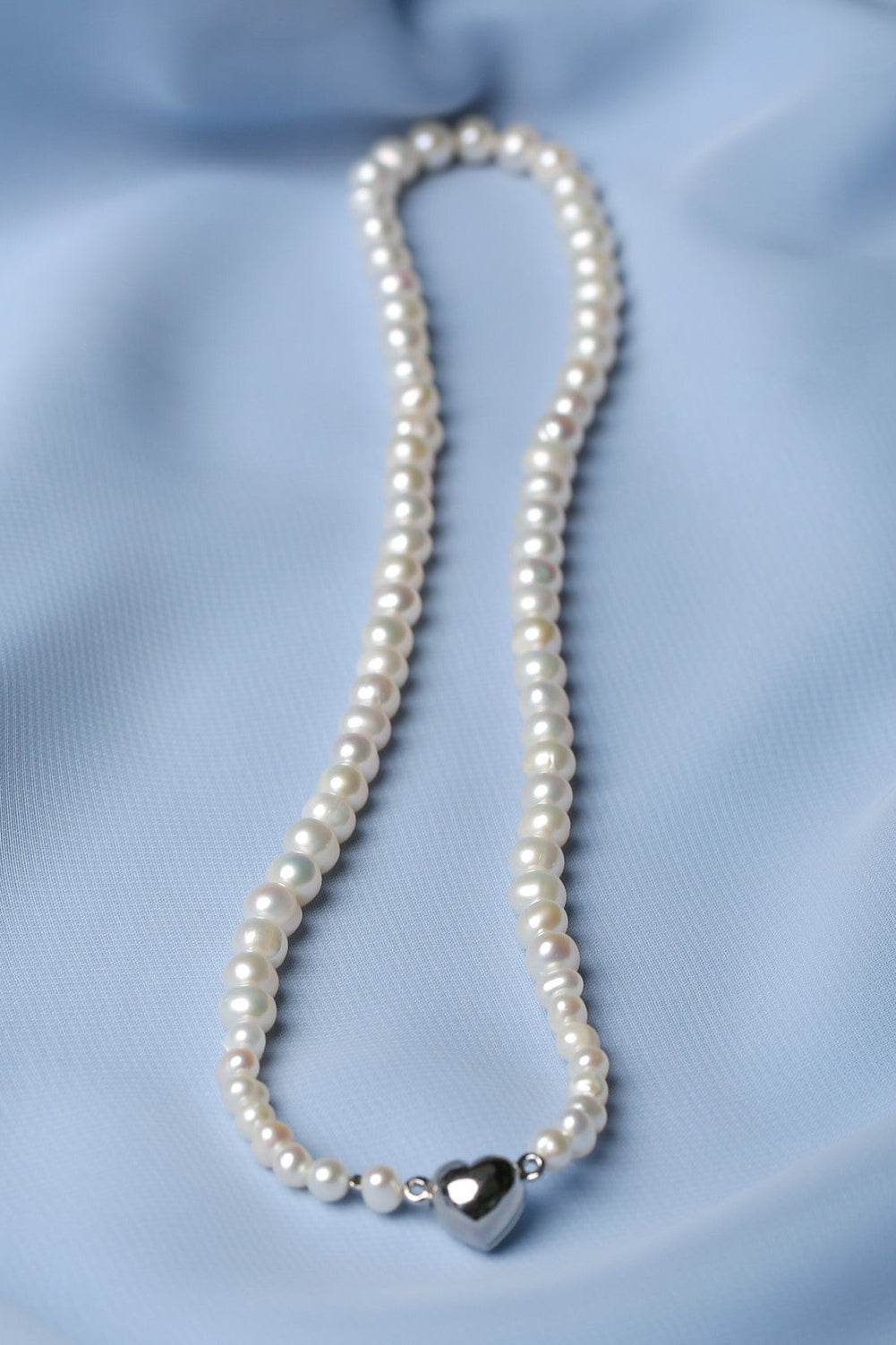 A strand of white pearl-like beads with a polished silver heart charm and small silver spacer beads, laid on soft light blue fabric.