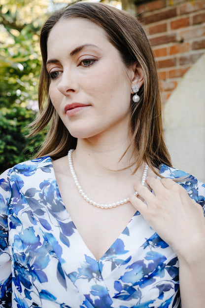 A woman wearing a blue and white floral print wrap dress and a pearl necklace, posing outdoors in front of a brick wall.