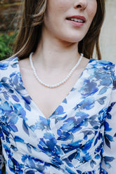 A woman wearing a blue and white floral print wrap dress with three-quarter sleeves, styled with a pearl necklace and bracelet in an outdoor garden setting.