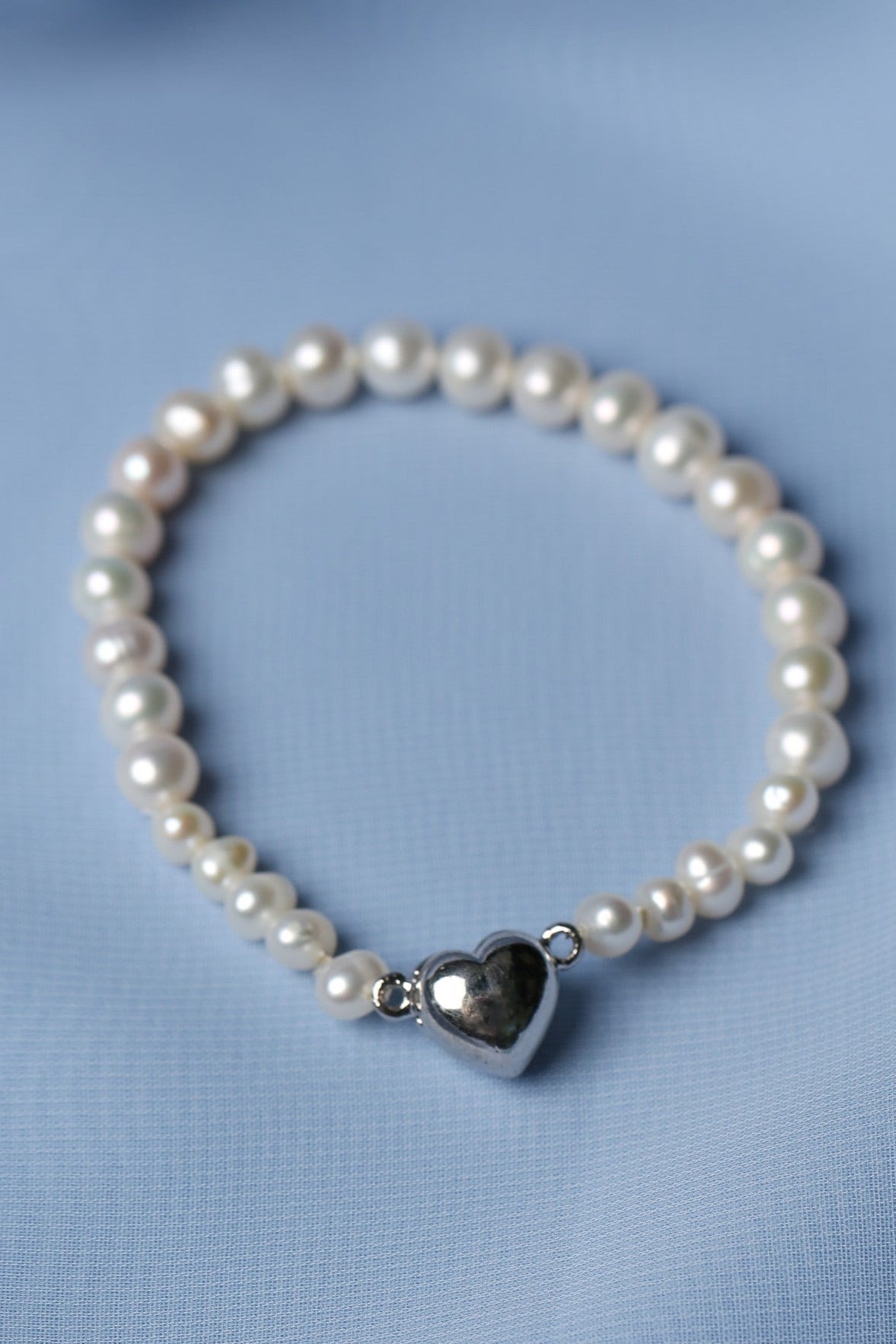 A beaded bracelet made of white freshwater pearls with a polished silver heart-shaped charm, displayed on a soft light blue fabric background.