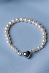 A beaded bracelet made of white freshwater pearls with a polished silver heart-shaped charm, displayed on a soft light blue fabric background.