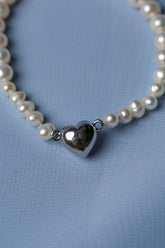 A close-up of a pearl strand necklace with a polished silver heart-shaped pendant, resting on a textured light blue fabric background.