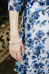 A close-up lifestyle shot of a woman wearing a white pearl bracelet with a small silver heart charm, paired with a blue and white floral dress.