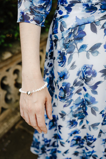 A close-up lifestyle shot of a woman wearing a white pearl bracelet with a small silver heart charm, paired with a blue and white floral dress.