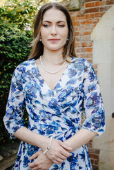 A woman wearing a blue and white floral print wrap dress, standing outdoors in front of a brick wall and stone archway.