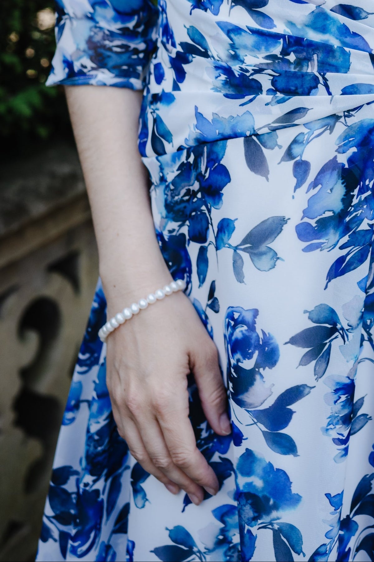 A woman wearing a blue and white floral print wrap dress with three-quarter sleeves, styled with a pearl necklace and bracelet in an outdoor garden setting.