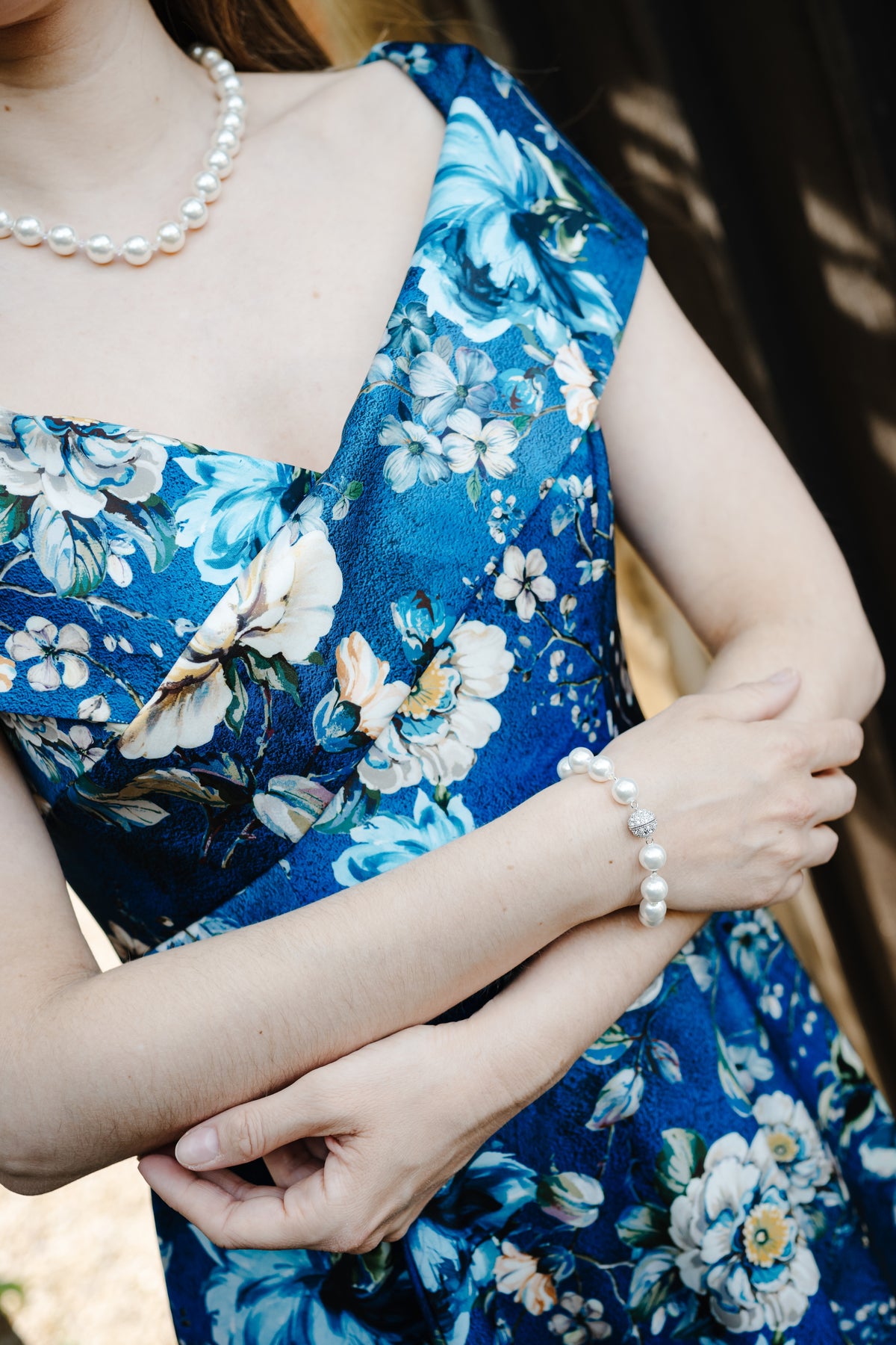 A close-up lifestyle shot of a woman wearing a blue floral dress with white and yellow flowers, accessorized with a pearl necklace and a pearl bracelet.
