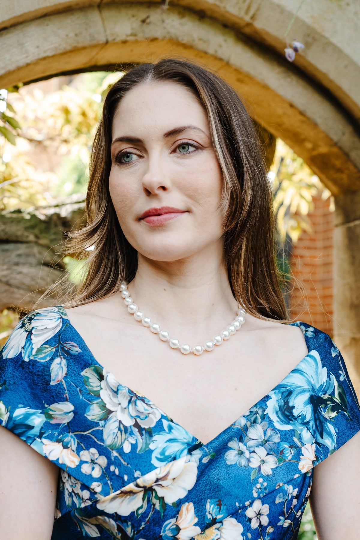 A woman wearing a blue floral dress with white and light blue flowers, styled with a pearl necklace and bracelet, standing in front of a stone archway.