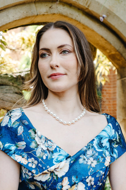 A woman wearing a blue floral dress with white and light blue flowers, styled with a pearl necklace and bracelet, standing in front of a stone archway.