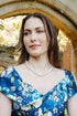 A woman wearing a blue floral dress with white and light blue flowers, styled with a pearl necklace and bracelet, standing in front of a stone archway.