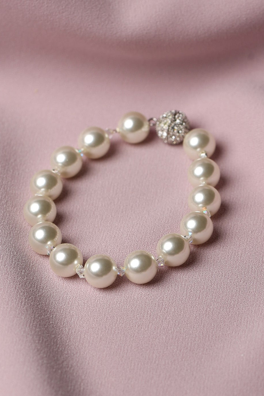A pearl beaded bracelet with a silver-toned rhinestone-embellished clasp, displayed on a soft pink satin fabric background.