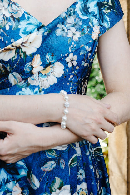 A woman wearing a blue floral dress with white and light blue flowers, styled with a pearl necklace and bracelet, standing in front of a stone archway.