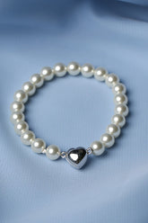 A beaded bracelet with white pearl-like beads and a shiny silver heart charm, displayed on a soft, draped light blue fabric background.