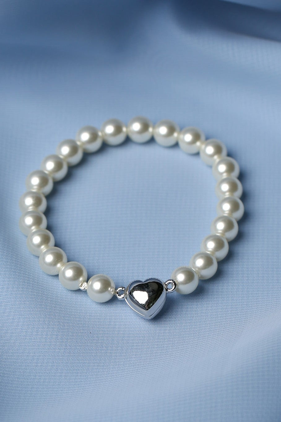 A beaded bracelet with white pearl-like beads and a shiny silver heart charm, displayed on a soft, draped light blue fabric background.