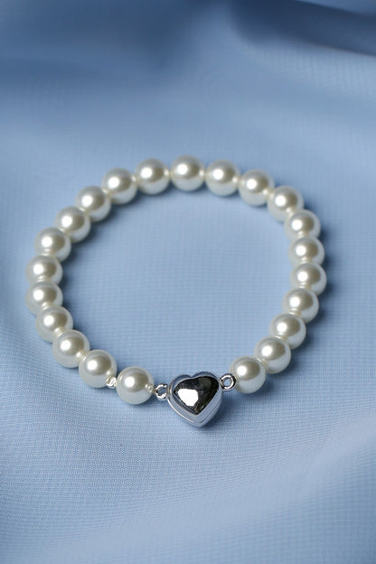 A beaded bracelet with white pearl-like beads and a shiny silver heart charm, displayed on a soft, draped light blue fabric background.
