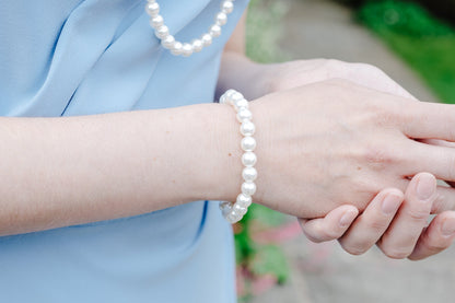 A close-up lifestyle shot of a person wearing a white pearl beaded bracelet on one wrist, paired with a light blue dress and a matching pearl necklace. The background is a softly blurred outdoor garden path.