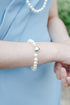 A close-up of a person wearing a white pearl bracelet with a silver heart charm, paired with a light blue dress and a matching pearl necklace, with a blurred outdoor garden background.