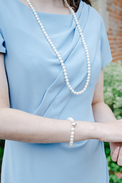 A woman wearing a light blue dress, a long pearl necklace, and a matching pearl bracelet with a small silver charm, standing outdoors in front of blurred greenery and a brick wall.