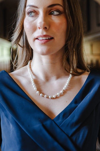 A woman with brown hair wearing a blue draped top and a pearl necklace, photographed in a softly lit indoor setting with an arched doorway in the background.