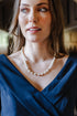 A woman with brown hair wearing a blue draped top and a pearl necklace, photographed in a softly lit indoor setting with an arched doorway in the background.