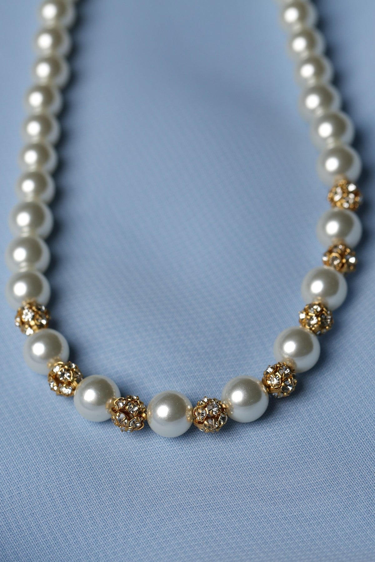 A pearl necklace with gold floral accents, displayed on a soft light blue fabric background.