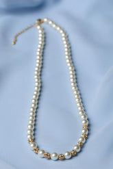 A pearl necklace with gold floral accents and a gold clasp, displayed on a soft light blue fabric background.