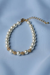 A pearl beaded bracelet with gold-toned decorative accents and a gold-toned extender chain, displayed on a soft light blue fabric background.