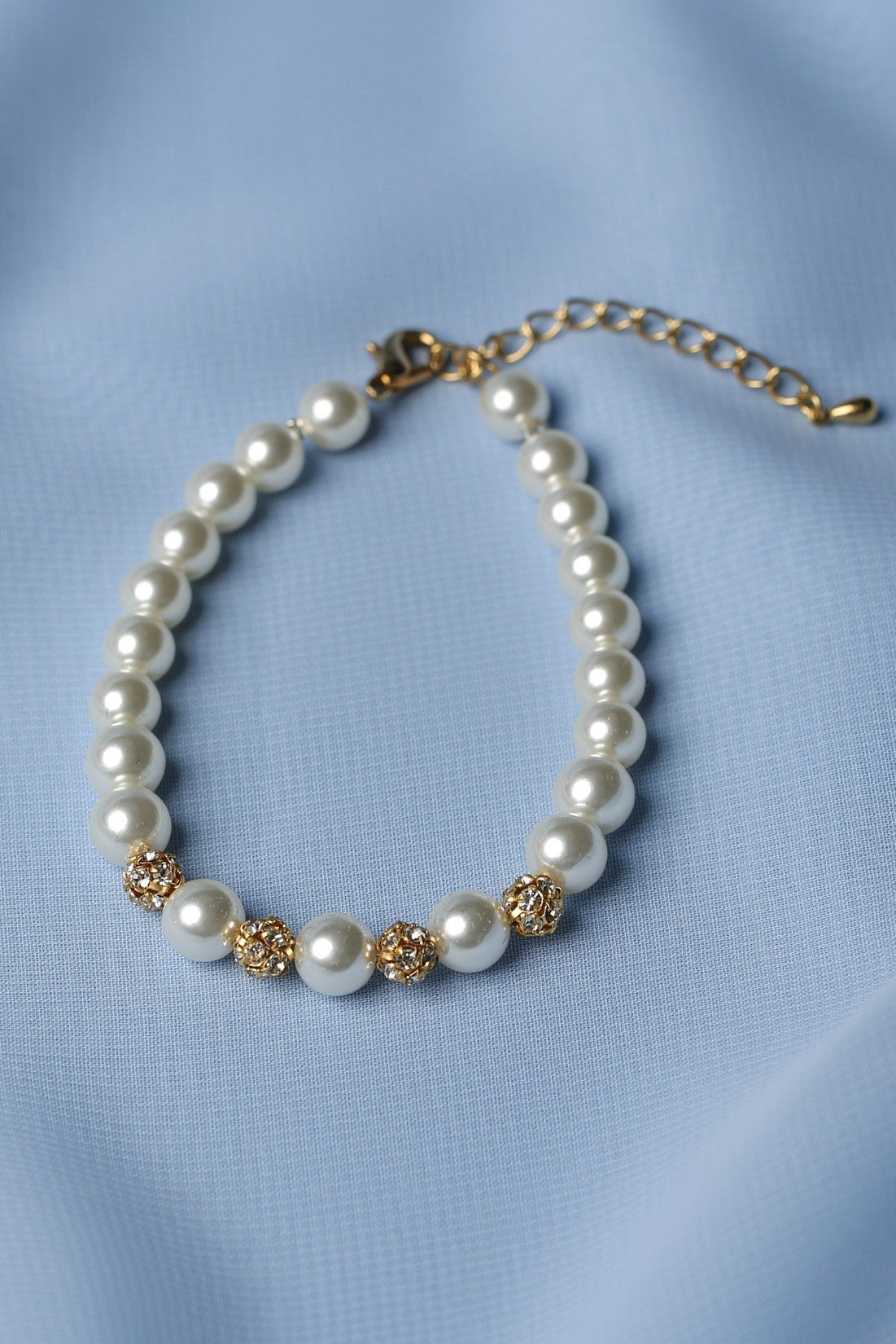 A pearl beaded bracelet with gold-toned decorative accents and a gold-toned extender chain, displayed on a soft light blue fabric background.