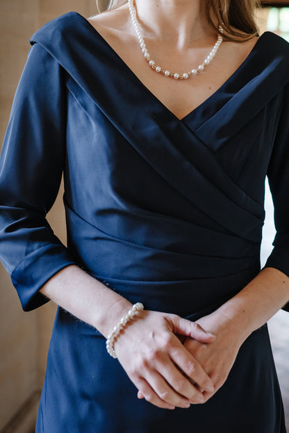 A woman wearing a navy blue wrap-style dress with three-quarter sleeves and a pearl necklace, standing in a softly lit indoor setting with stone arches and a blurred background.