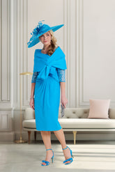Woman in a blue dress and hat standing in a stylish room with a sofa and decorative pillows.